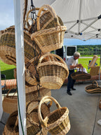Several natural woven wicker baskets with handles are displayed on a white rack outdoors under a canopy tent, with a person and other items visible in the background.