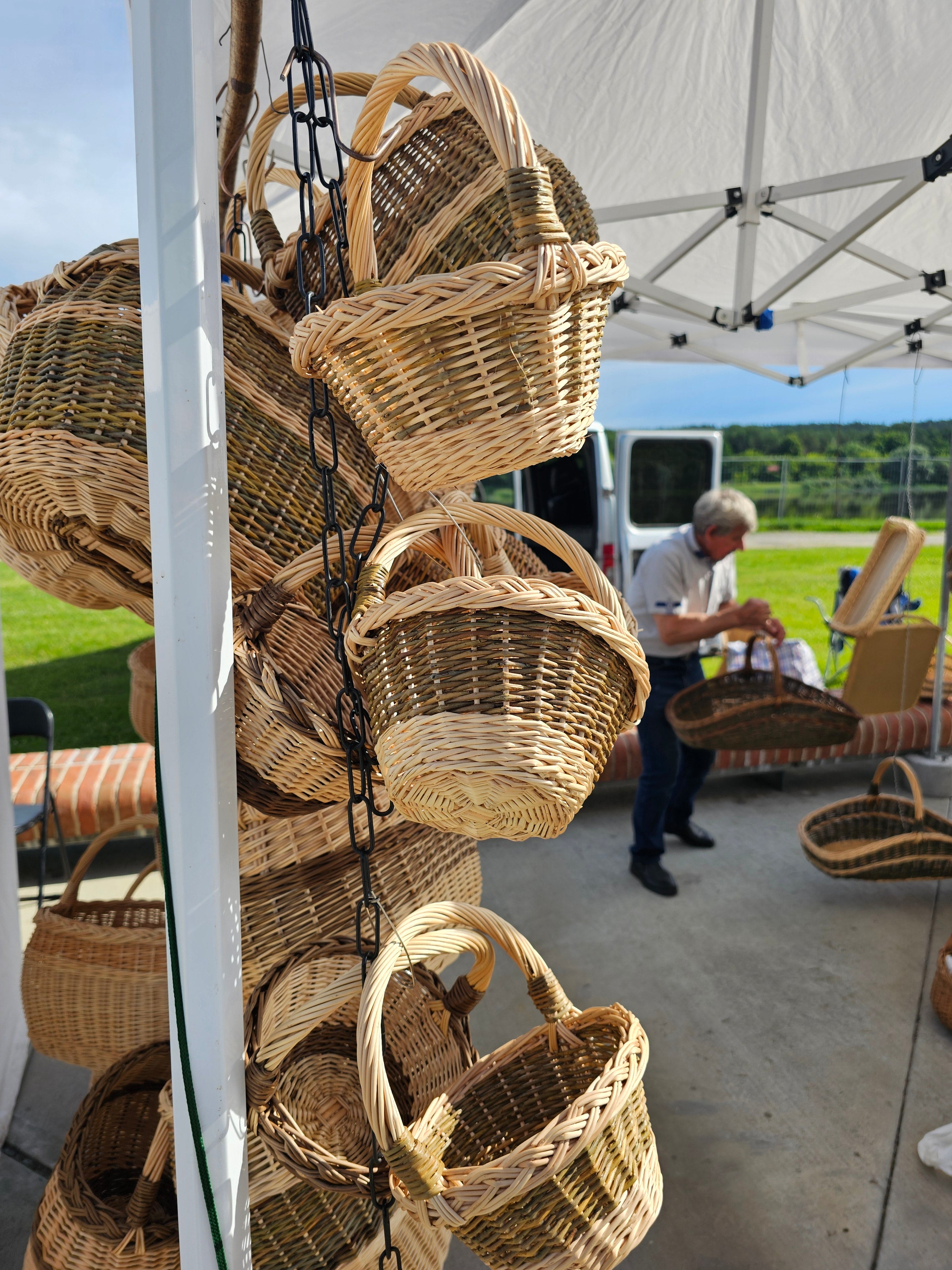 Several natural woven wicker baskets with handles are displayed on a white rack outdoors under a canopy tent, with a person and other items visible in the background.